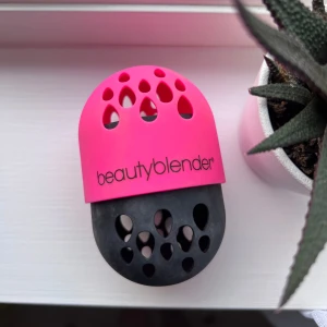Förvaring till Beautyblender  - Säljer ett Beautyblender förvaringsetui i silikon, perfekt för att skydda och torka din beautyblender. Etuiet är tvådelat i stark rosa och svart med droppformade ventilationshål och logga på framsidan. Smidig att ta med i väskan eller ha på sminkbordet. Har tvättat av så gott det går!