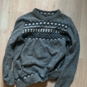 Grå stickad tröja med mönster - Mysig grå stickad tröja med klassiskt nordiskt mönster i vitt och mörkgrått över bröstet och ärmsluten. Tröjan har rund halsringning och långa ärmar, perfekt för kyliga dagar.