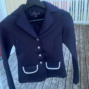 Mörkblå ridkavaj från CRW Exclusive - Snygg mörkblå ridkavaj från CRW Exclusive med silvriga knappar och vita detaljer vid fickorna. Klassisk krage och figurnära passform, perfekt för dig som vill sticka ut på tävling eller träning. Långärmad och stilren design som ger en proffsig look. Frakten tar köparen. 