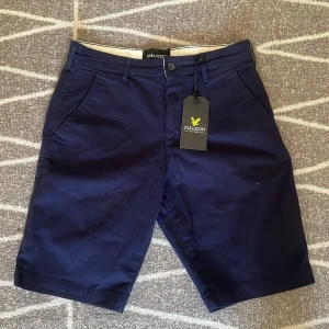 Mörkblå chinoshorts från Lyle & Scott - Snygga mörkblå chinoshorts från Lyle & Scott i klassisk modell med raka ben och sidfickor. Shortsen har midwaist-passform och är tillverkade i bomull, perfekta för sommardagar. Stilren design med knapp och dragkedja framtill.