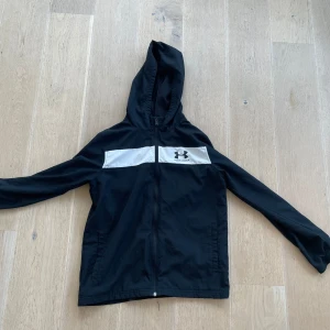 Svart vindjacka från Under Armour - Svart vindjacka med vit panel över bröstet och Under Armour-logga. Jackan har huva, dragkedja framtill och långa ärmar. Perfekt för blåsiga dagar och sportiga outfits.