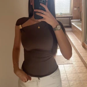 Brun one shoulder topp med gulddetalj - Säljer en tight brun topp med one shoulder-design och snygg guldfärgad detalj vid axeln. Toppen är ärmlös och har en slimmad passform som framhäver figuren. Perfekt för dig som gillar trendiga och unika plagg. Från zara