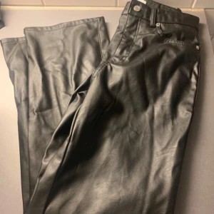 Svarta byxor i skinnimitation - Snygga svarta byxor i skinnimitation med klassisk femficksmodell och raka ben. Byxorna har bälteshällor, knapp och dragkedja framtill. Perfekta för dig som vill ha en edgy look med lite attityd.