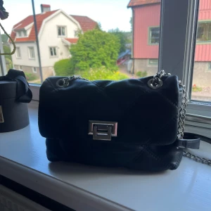 Svart stradivarius väska med kedja - Snygg svart axelväska i quiltad design med silverfärgad kedjerem och metallspänne framtill. Väskan har både handtag och axelrem, samt ett innerfack med dragkedja. Perfekt storlek för det viktigaste och ger en cool vibe till din outfit.