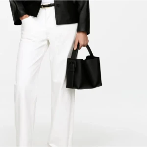 Svart handväska i skinn med spänne - Stilren svart handväska i skinn med korta handtag och snyggt spänne på sidan. Väskan har en boxig form och minimalistisk design som passar till många outfits. Perfekt storlek för det viktigaste du behöver ha med dig.