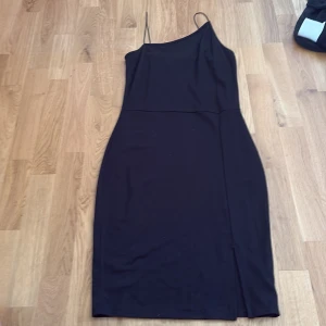 Svart oneshoulder klänning från NA-KD - Snygg svart klänning från NA-KD med oneshoulder och smala axelband. Modellen är kroppsnära med en liten slits nertill och har en stilren, minimalistisk look. Perfekt för dig som gillar enkel men trendig stil. Liten i storlek, passar någon som är s/M