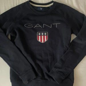 Mörkblå sweatshirt från Gant XS - Snygg mörkblå sweatshirt från Gant med broderad logga och sköld med stjärnor och ränder på bröstet. Tröjan har rund halsringning och långa ärmar. Perfekt för dig som gillar klassisk och sportig stil.