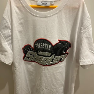 Vit Trapstar t-shirt London Shooters - Vit t-shirt från Trapstar med stort broderat London Shooters-motiv och panter på bröstet. Klassisk rund hals, korta ärmar med mesh-detaljer på axlarna. Skön passform och streetwear-vibe, perfekt för dig som gillar statement-plagg.