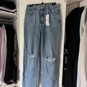 Ljusblå ripped jeans med raka ben - Säljer ett par ljusblå jeans med raka ben och slitna hål över knäna. Klassisk femficksmodell i denim med normal passform och medelhög midja. Perfekta för en avslappnad och trendig look. Önskar du veta exakta mått? Tveka inte att höra av dig!
