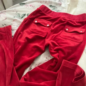 Röda velourbyxor Juicy Couture S - Röda mjukisbyxor från Juicy Couture i mjuk velour med bred resår i midjan och fickor med lock och knappar bak. Klassisk logga broderad framtill och coola detaljer. Perfekta för en chill och trendig look.