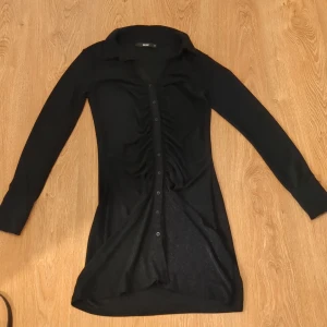 Svart skjortklänning från Bik Bok stl. M - Svart kort skjortklänning från Bik Bok med långa ärmar, krage och knappar hela vägen framtill. Snygg rynkdetalj längs knappraden. Aldrig använt 