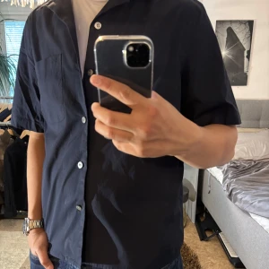 Mörkblå skjorta från Calvin Klein - Snygg mörkblå skjorta från Calvin Klein i relaxed fit. Skjortan har korta ärmar, klassisk krage, bröstficka med diskret 'ck'-brodyr och knäppning framtill. Tillverkad i mjuk bomull, perfekt för en clean och stilren look.