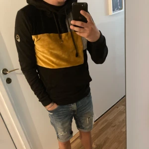 Vichy kofta - Säljer en Vichy hoodie med bred gul panel över bröstet och snörning i huvan. Tröjan har lång ärm, känguruficka och en liten logga på ärmen. Perfekt för dig som gillar streetstyle och vill sticka ut med färg.