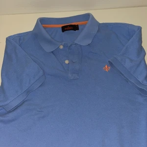 Blå pikétröja från Morris, storlek M - Snygg blå pikétröja från Morris med klassisk krage och knäppning framtill. Tröjan har korta ärmar och en orange broderad logga på bröstet. Perfekt för en stilren och avslappnad look. Tillverkad i bomull för skön känsla.