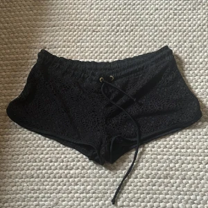 Svarta lågmidjade spetsiga shorts med snörning - Svarta shorts med elastisk midja och snörning framtill. Utsidan är täckt av ett snyggt spetsmönster som ger en cool och feminin vibe. Perfekta för varma dagar eller som strandplagg. Mjukt innerfoder för extra komfort.