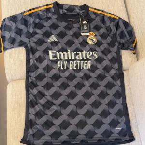 Real Madrid Adidas fotbollströja svart - Snygg svart Real Madrid fotbollströja från Adidas med grått grafiskt mönster på framsidan och ärmarna. Tröjan har gula detaljer på axlarna och klubbmärke på bröstet. Tillverkad i lätt och ventilerande material, perfekt för träning eller match.