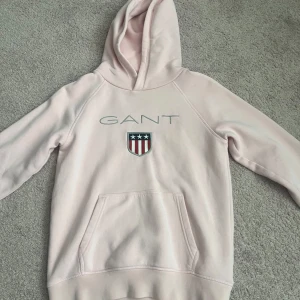 Ljusrosa hoodie från GANT med logga - Ljusrosa hoodie från GANT med broderad logga och sköld på bröstet. Tröjan har huva, magficka och långa ärmar. Mjuk insida och klassisk passform, perfekt för chill dagar. Snygg detalj med randigt band på insidan av kragen. Storlek 146/152 men är som en xxs/xs. Använd fåtal gånger. Säljer pågrund av att denna tröja är för liten för mig. Skriv till mig innan ni köper tröjan.