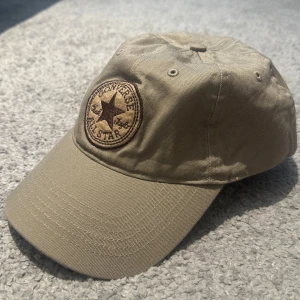 Beige keps från Converse All Star - Snygg beige keps från Converse All Star med broderad logga framtill. Justerbar rem baktill för perfekt passform. Tillverkad i 100% bomull, vilket gör den både bekväm och lätt att bära. Perfekt accessoar för en avslappnad streetstyle.