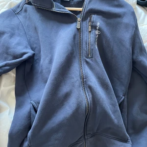 Blå zipjacka från Sail Racing - Blå zipjacka från Sail Racing med dragkedja framtill och en extra bröstficka med dragkedja. Jackan har hög krage och två sidofickor, perfekt för en chill och sportig stil. Broderad logga på bröstet ger en clean look.