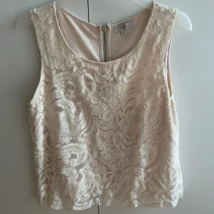 Beige spetslinne med dragkedja - Supersött beige linne  med vacker spets och blommigt mönster. Toppen är ärmlös och har en synlig dragkedja i ryggen. Perfekt att styla med jeans eller kjol för en romantisk vibe