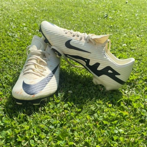 Nike Air Zoom fotbollsskor gul/svart - Säljer ett par Nike Air Zoom fotbollsskor i vitt med svarta detaljer och stora swoosh-loggor. Skorna har snörning och är tillverkade i syntetmaterial för lätt känsla på planen. Perfekta för dig som vill sticka ut med stilrena och snabba skor. Och det som är lite orange som syns är bara lite smuts som man säkert kan ta bort, inget fel med dom och rekommenderar dom.