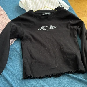 Svart långärmad tröja med hjärtdetalj - Svart långärmad tröja från FB Sister i storlek XS. Framtill finns ett broderat hjärta med vingar i vitt. Tröjan har en croppad passform med vågig, rå kant nertill för en edgy look. Perfekt till jeans eller kjol för en cool vardagsstil.