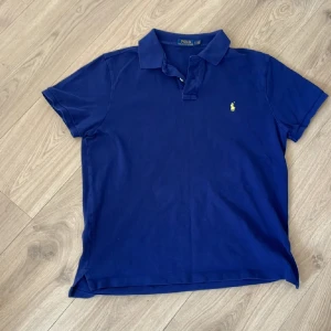 Marinblå pikétröja från Polo Ralph Lauren - Klassisk marinblå pikétröja från Polo Ralph Lauren med gul broderad logga på bröstet. Tröjan har korta ärmar, krage och knappar vid halsen. Tillverkad i bomull för en skön och avslappnad känsla.
