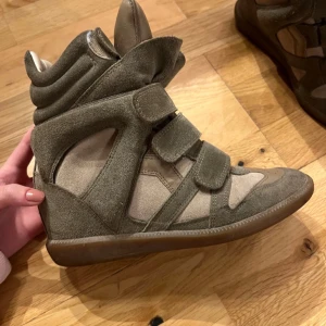 Isabel Marant wedge sneakers mocka - Coola Isabel Marant sneakers i olivgrön och beige mocka med högt skaft och dolda kilklackar. Skorna har kardborreband, rund tå och detaljer i skinn. Perfekta för dig som vill ha en sportig men edgy look.