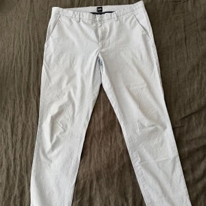 Randiga sommarchinos från BOSS i ljusblått - Snygga ljusblå och vita randiga chinos från BOSS med stretch. Klassisk passform med fickor i sidorna, bälteshällor och dragkedja. Perfekta för en clean och stilren look, med diskret metallogo bak. Tunt och skönt bomullsmaterial perfekt för sommaren.