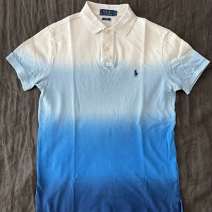 Blåvit pikétröja från Polo Ralph Lauren - Säljer en snygg pikétröja från Polo Ralph Lauren i storlek S. Tröjan har en cool ombre-effekt från vitt upptill till blått nedtill, klassisk krage och två knappar. Ikoniska Polo-loggan broderad på bröstet. Perfekt för dig som gillar stilrena och fräscha plagg.
