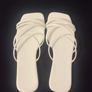 Vita sandaler med tunna remmar - Stilrena vita sandaler med fyrkantig tå och flera tunna remmar som korsar foten. Platt sula och minimalistisk design, perfekta för en trendig look. Snygga att matcha med både jeans och klänning under varma dagar.