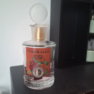 Monotheme Pomegranate Eau de Toilette - Fräsch parfym från Monotheme med doft av granatäpple. Flaskan är cylindrisk i klart glas med en transparent rund kork och gulddetaljer vid halsen. Etiketten är röd med illustration av granatäpple och gröna blad. Innehållet är ljusrosa. 100 ml från början. 