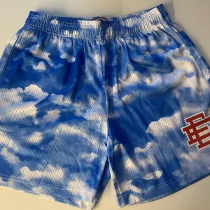 Blå molnmönstrade meshshorts EE M - Snygga meshshorts med blått och vitt molnmönster och röd EE-logga på ena benet. Shortsen har elastisk midja och är perfekta för en chill och sportig look. Lätta och luftiga, passar grymt till sommaren.