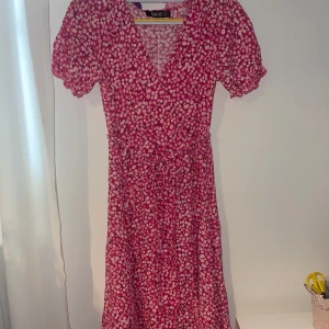 Rosa blommig långklänning från Mico - Supersöt kortklänning i rosa med små vita blommor från Mico. Klänningen har v-ringning, korta puffärmar och ett knytband i midjan som ger en snygg siluett. Perfekt för varma dagar när du vill känna dig extra fin.