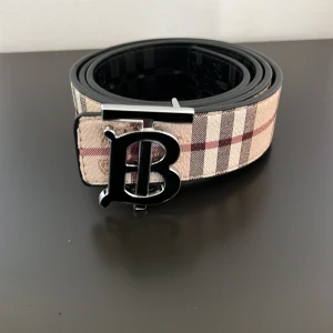 Rutig Burberry bälte med B-spänne - Snyggt bälte från Burberry med klassiskt rutigt mönster i beige, svart och röd. Bältet har ett stort metallspänne med ett stilrent B i silverfärg. Tillverkat i skinn med exklusiv känsla och passar perfekt för att lyfta din outfit.