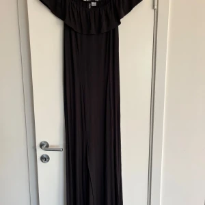 Svart långklänning från Divided - Svart långklänning från Divided med offshoulder-modell och bred volang upptill. Klänningen är rak i modellen och har en mjuk, skön känsla. Perfekt för dig som gillar en enkel men snygg stil.