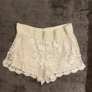 Vita virkade spets-shorts - Superfina vita shorts med bred resår i midjan och detaljerad virkad spets över hela plagget. Blommiga mönster och vågig kant nedtill ger en romantisk och somrig vibe. Perfekta för varma dagar och festivaler.