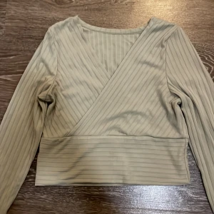 olivgrön omlottopp från SHEIN - Snygg Oliv tonad omlottopp från SHEIN i ribbad polyester. Toppen har långa ärmar, croppad passform och ett stilrent randigt mönster, toppen är lagomt stretchig. Perfekt för dig som gillar en enkel men trendig look med lite edge. 