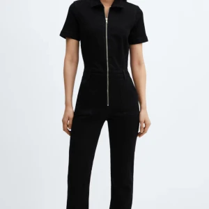 Svart jumpsuit med dragkedja - Snygg svart jumpsuit med korta ärmar och rak passform. Dragkedja framtill som går hela vägen upp till kragen. Tillverkad i ett mjukt bomullsmaterial som ger en clean och stilren look.