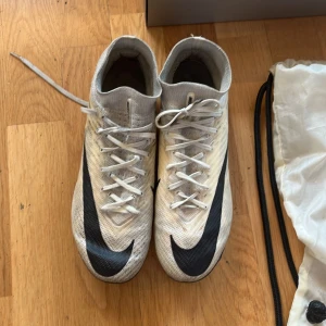 Nike Mercurial Air Zoom fotbollsskor Elite - Nike Mercurial Air Zoom fotbollsskor i vitt med svarta detaljer och stor Swoosh-logga. Skorna har snörning, platt sula med dobbar och är tillverkade i syntetmaterial för lätt känsla på planen. Medföljer originalpåse med Zoom Mercurial-tryck (inte original boxen till). Skorna köptes för 3400 kr och är använda i 4 månader av min son som är 13 år. 