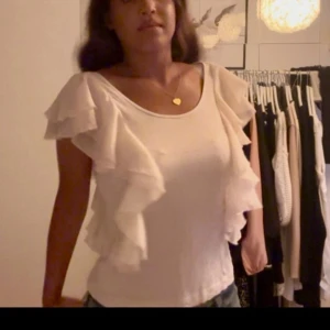 Ljus rosa/vit blus med volangärmar - Supersöt vit blus med stora volanger längs ärmarna och axlarna. Blusen har rund halsringning och är tillverkad i ett lätt, flowy tyg som ger en romantisk vibe. Perfekt att styla med jeans eller kjol för en trendig look.