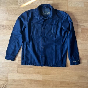 Mörkblå overshirt från D'once De La Sense - Stilren mörkblå overshirt från D'once De La Sense i ull. Klassisk krage, två stora bröstfickor och knäppning framtill. Broderad gul logga på ärmen ger en unik touch. Perfekt lager-på-lager-plagg för kyligare dagar. Med den nyaste modellen. 