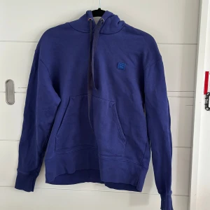 Blå hoodie från Acne Studios - Säljer en blå hoodie från Acne Studios med klassisk smiley-patch på bröstet. Tröjan har huva med snörnin och långa ärmar. Sällan använd har en liten fläck på bröstet men svår att se
