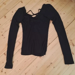Svart långärmad topp från Monki XS - Svart långärmad topp från Monki i storlek XS. Toppen har en snygg v-ringning med tunna banddetaljer över bröstet och är figurnära i modellen. Perfekt att styla med jeans eller kjol för en cool look.