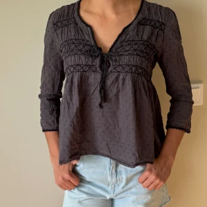 Odd molly blus - Supersöt svart blus från Odd Molly med v-ringning, knyt framtill och broderade detaljer över byst och ärmar. Lätt transparent tyg med diskret prickmönster och trekvartsärmar. Perfekt för dig som gillar boho-vibe och romantiska detaljer.