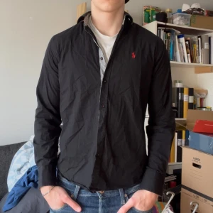 Svart skjorta från Ralph Lauren - Snygg svart skjorta från Ralph Lauren med röd broderad logga på bröstet. Skjortan har en slim fit passform och långa ärmar. Insidan av kragen har ett stilrent rutigt mönster. Perfekt för en stilren look.