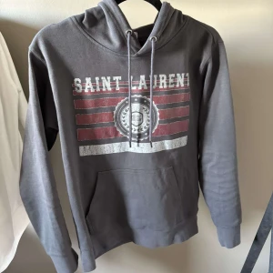 Grå Saint Laurent hoodie med tryck - Säljer nu en Saint Laurent Hoodie åt min vän i storlek M. Den är i nyskick och skulle säga den passar 175-185. Osäker om äkta köpt för 2300!