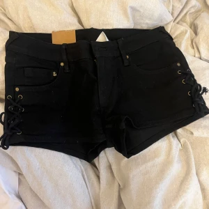Svarta jeansshorts med snörning H&M - Svarta, tighta jeansshorts från H&M med snygg snörningsdetalj i sidorna och klassiska fickor både fram och bak. Shortsen har låg midja och är gjorda i stretchig denim som sitter skönt. 