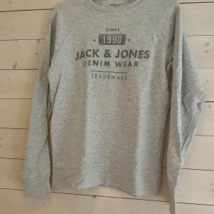 Grå sweatshirt från Jack & Jones - Snygg ljusgrå sweatshirt från Jack & Jones med tryck framtill. Tröjan har rund halsringning och långa ärmar. Perfekt basic-plagg med avslappnad passform och mjukt bomullsmaterial.