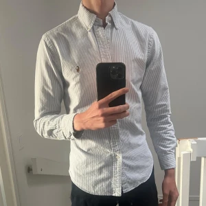 Randig skjorta från Polo Ralph Lauren - Säljer en slim fit skjorta från Polo Ralph Lauren i storlek S. Skjortan är vit med tunna blå ränder, har klassisk button-down krage, långa ärmar och broderad logga på bröstet. Tillverkad i bomull och perfekt för en clean och stilren look.